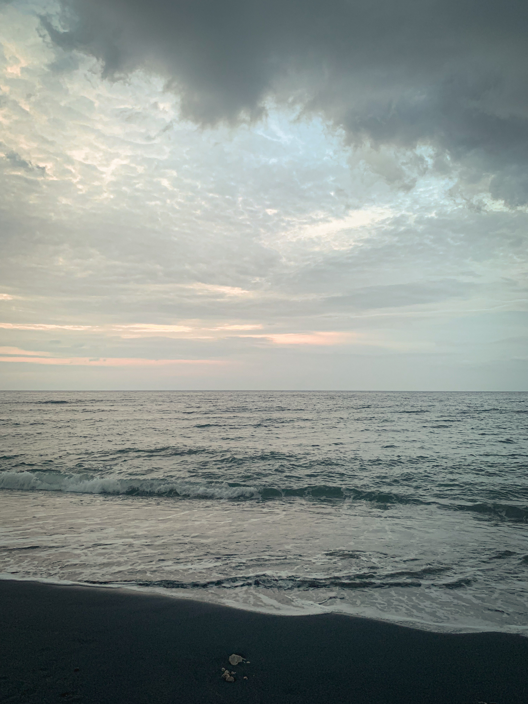 photo of the beach with a muted tone, with hazy grayish clouds across blue sky with faint pink and gold highlights, pewter clouds hovering over the top of the image, and silver-green water washing up onto black sand. With a small arrangement of stones or worn-away coral submerged in the sand.