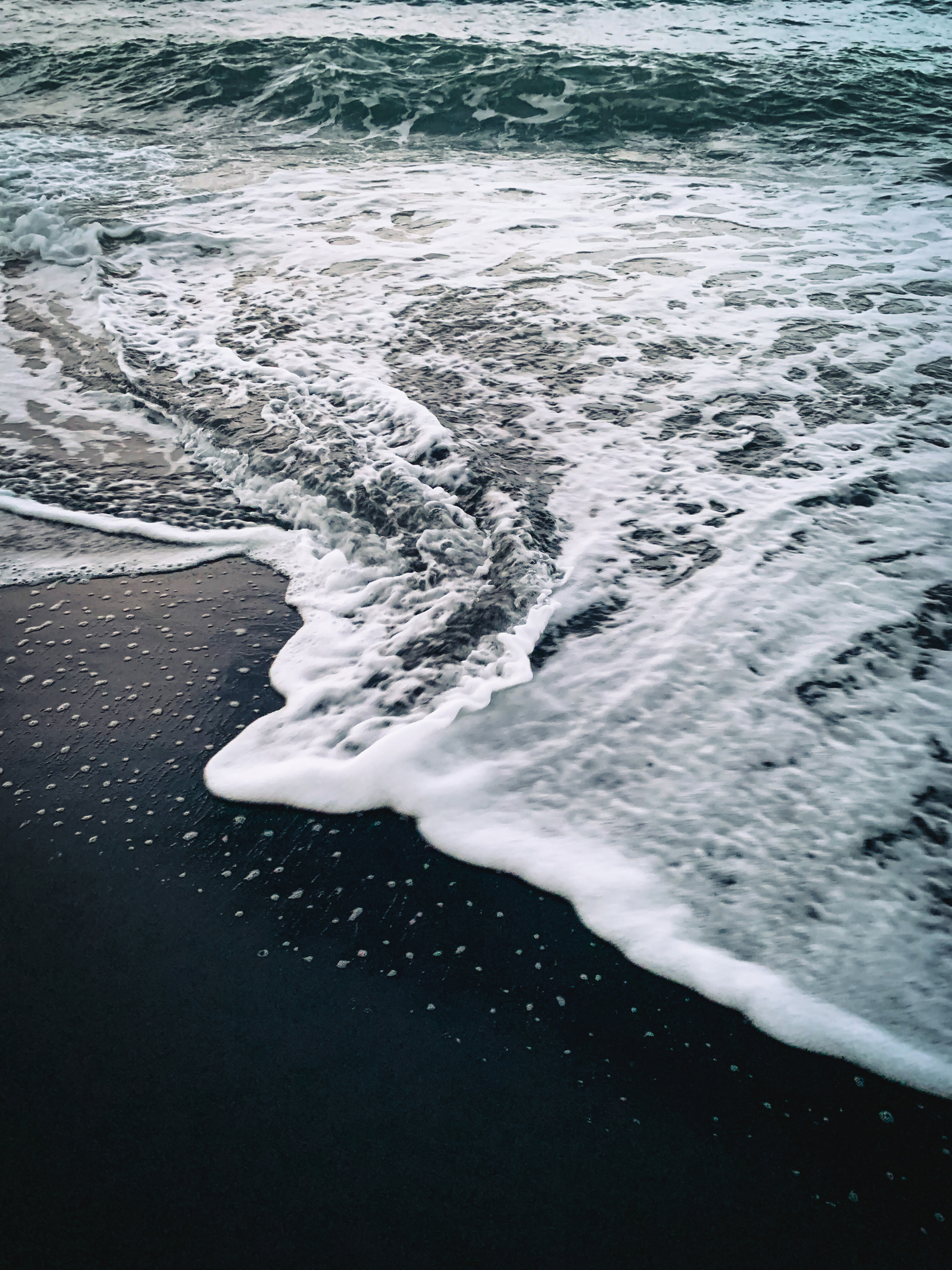 dramatic photo at a beach with deep black sand, a white frothy wave coiling across the image, leaving a stellar speckle of white froth scattered across sand, and sea-green surging water in the background.