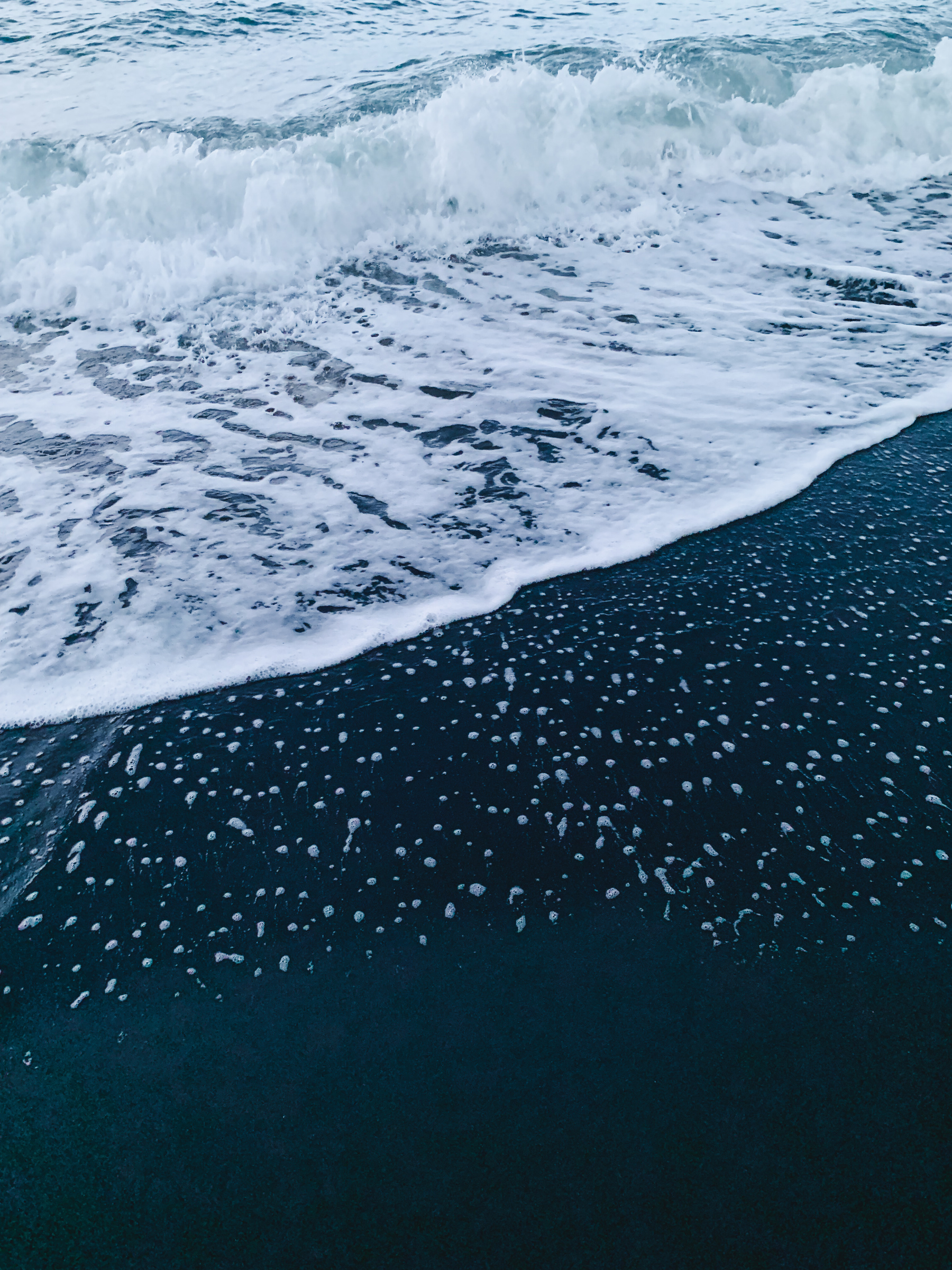 photo of water washing up on the beach, in a choppy-frothy wave of icy turquoise water skimming across nearly black-blue sand, leaving a dense speckle of frothy white bubbles.