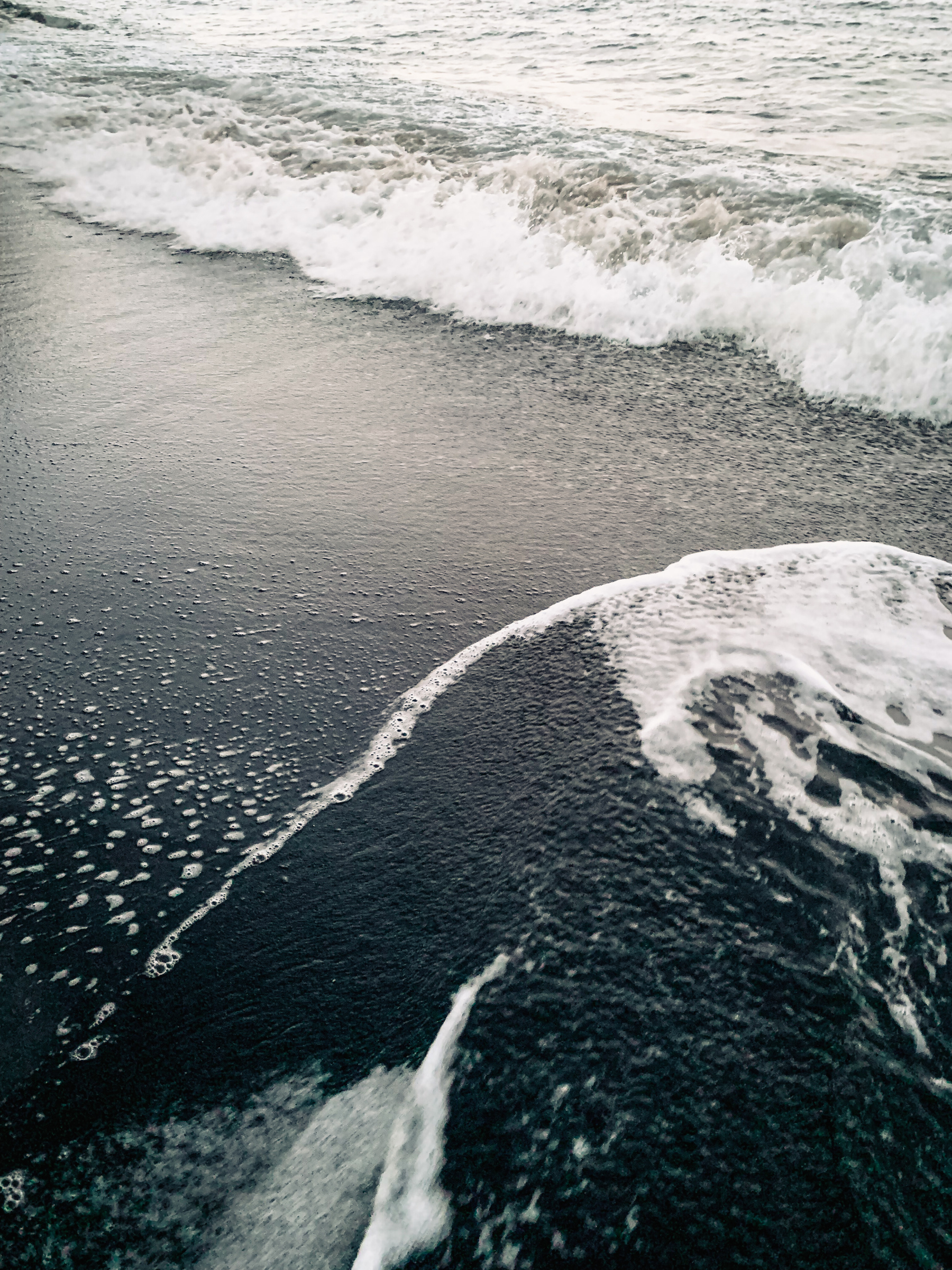 photo of water washing up on a black-sand beach in dynamic curvilinear motions, falling back into a frothy, on-coming wave, the surface of the water coarsened by falling rain, with white foam swirling over and speckling the dark sand, sometimes blurring into motion, and a warm yellowish, antique cast to the light.
