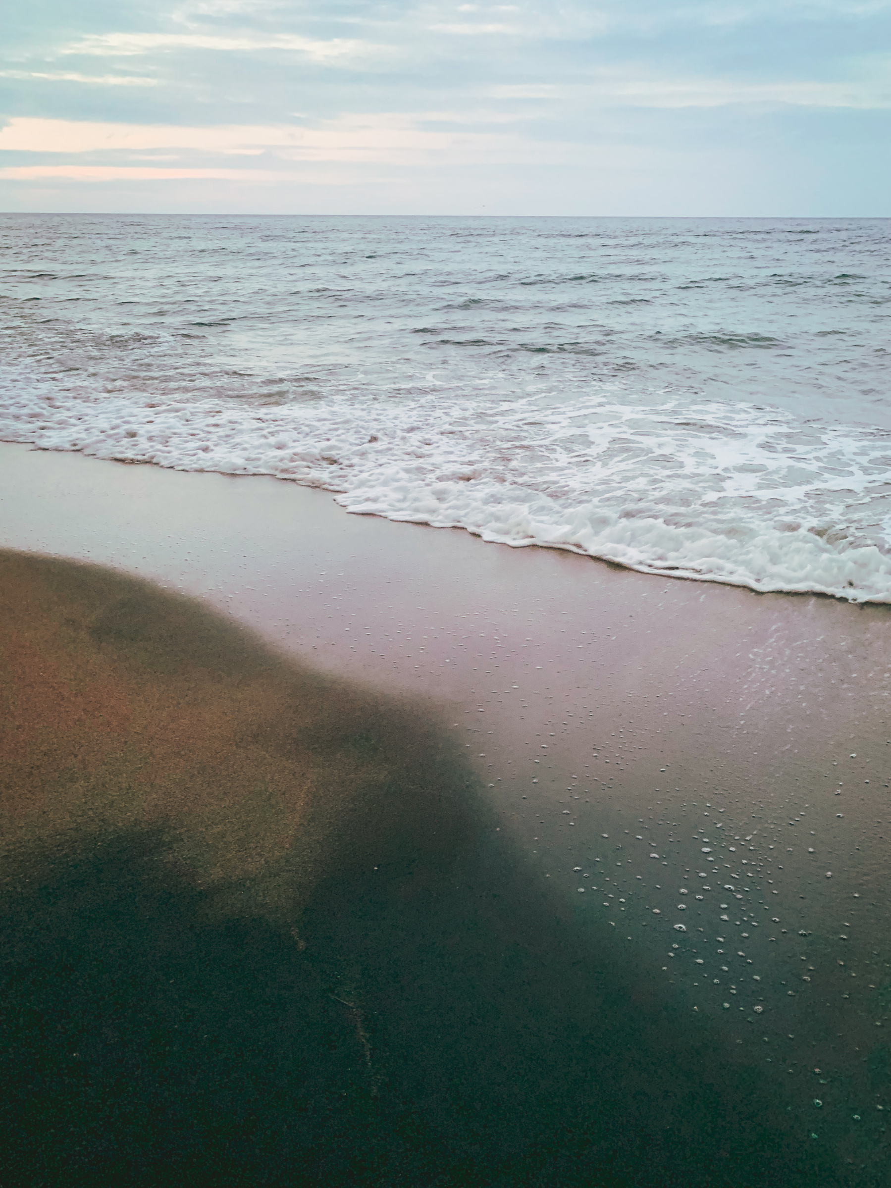 photo of a beach in the early morning with an eerie and soft tone, pale and fuzzy light in mostly silver, greenish, and gold-ish shades, a glowing froth of gentle surf, with some warmer rose reflected off black sand, blurred with a ghostly and fleeting yellowish-sienna shape, speckled with spots of foam