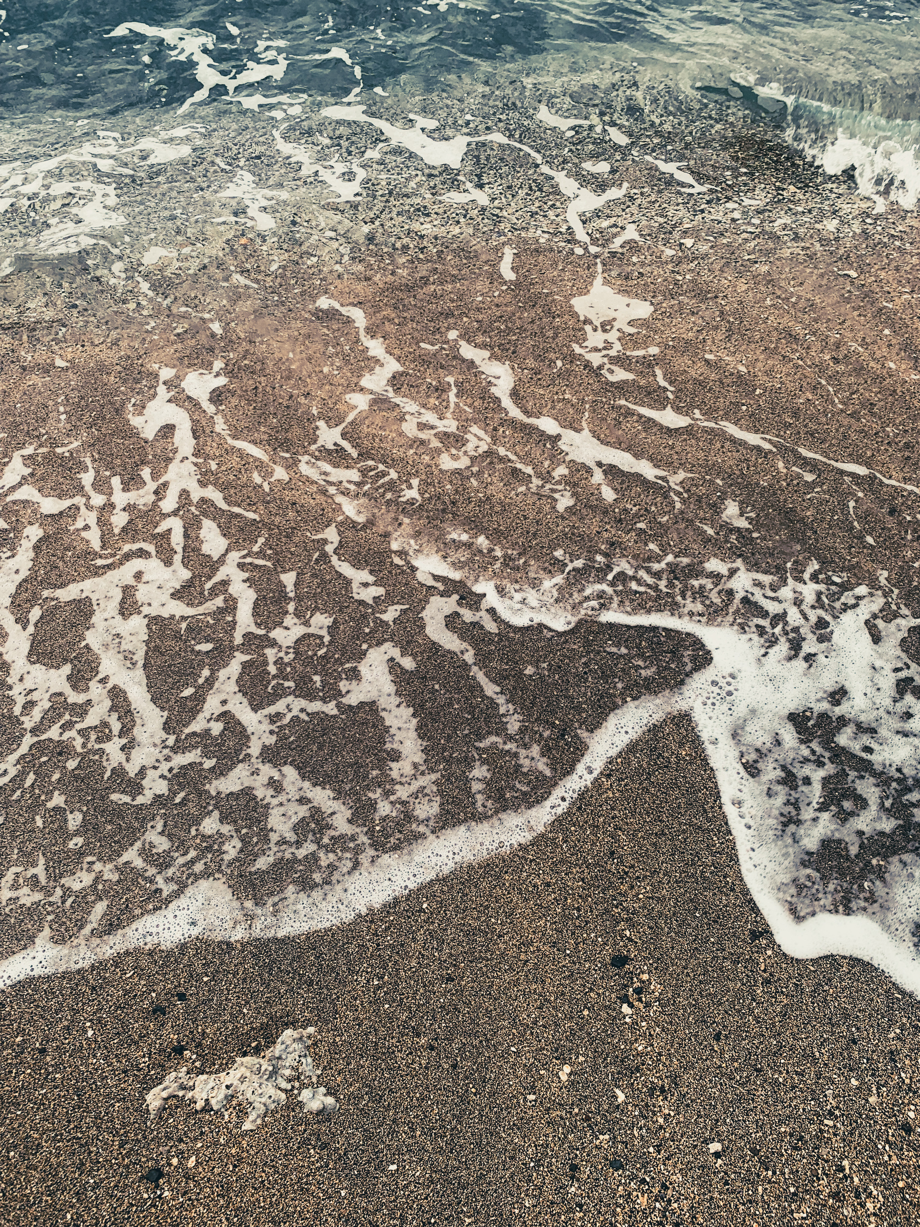 photo of surf at the beach, foamy and frothy translucent turquoise water with beige, golden, black speckled sand and gravel and a worn down piece of off-white coral submerged in the sand.