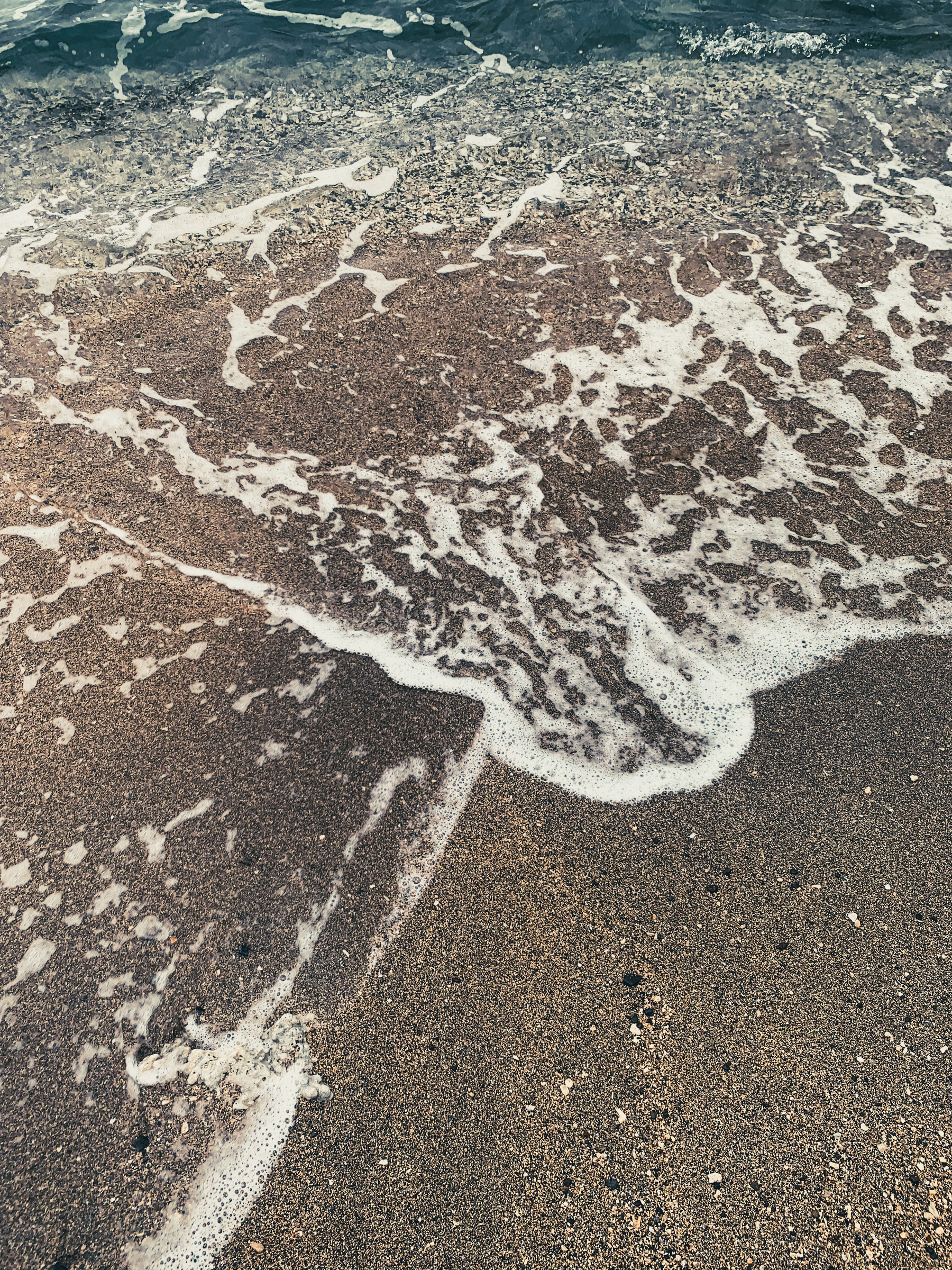 photo of turquoise blue water rushing over specked tan and black sand at the beach, spilling past its own boundary.