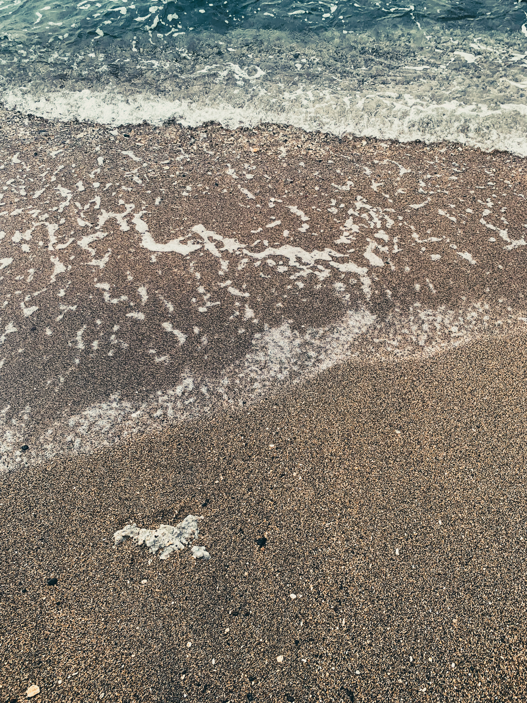 close-up photo of water moving over the sand on the beach, with jelly-like turquoise blue ripple on the top, cascading into a translucent layer of lattice frothy foam over the speckled tan and black sand, studded with polished bits of shells and coral.