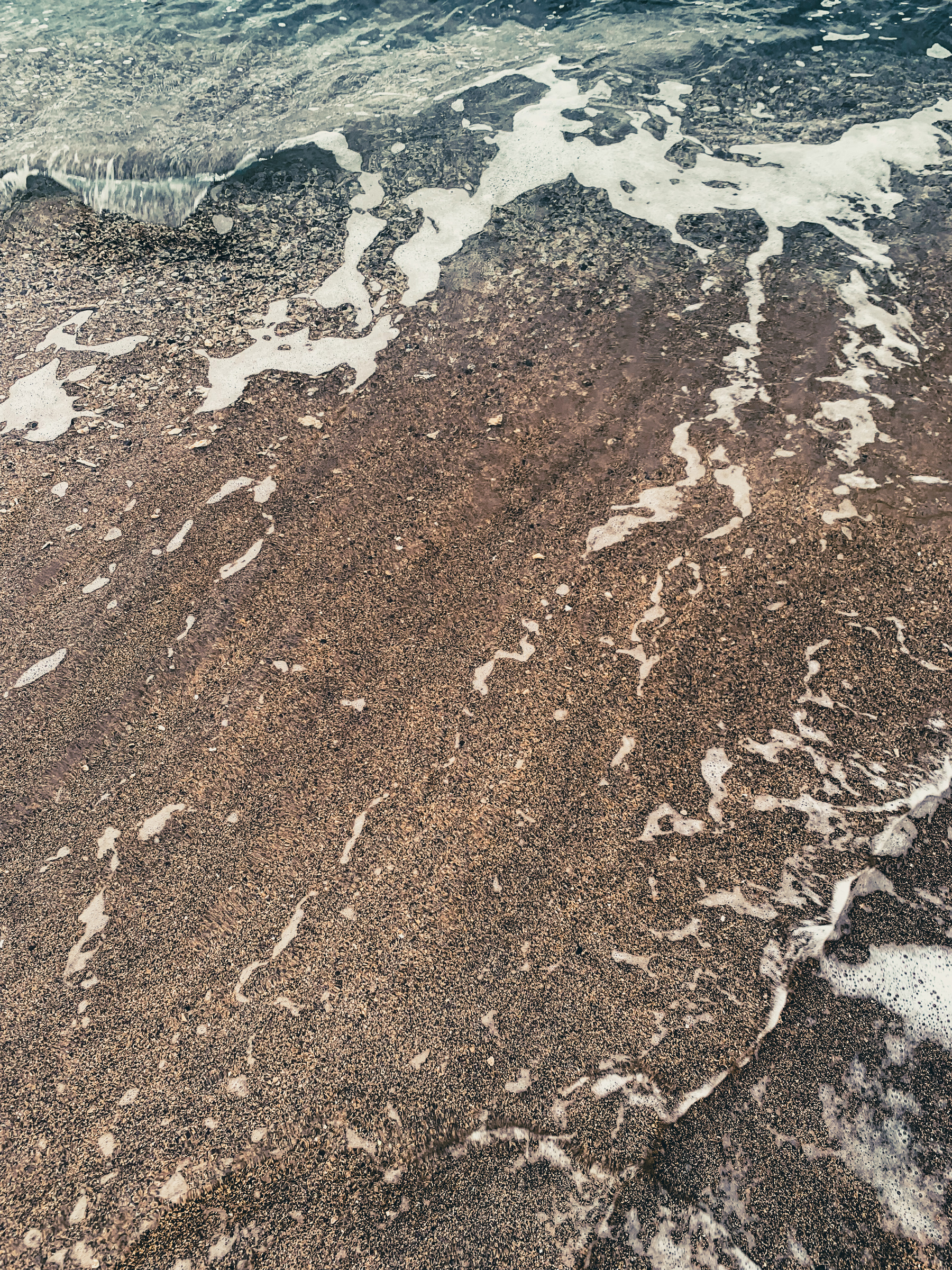 closer-up photo of a frothy wave at the beach, turquoise water transparently and completely having covered tan-brown and black speckled sand.