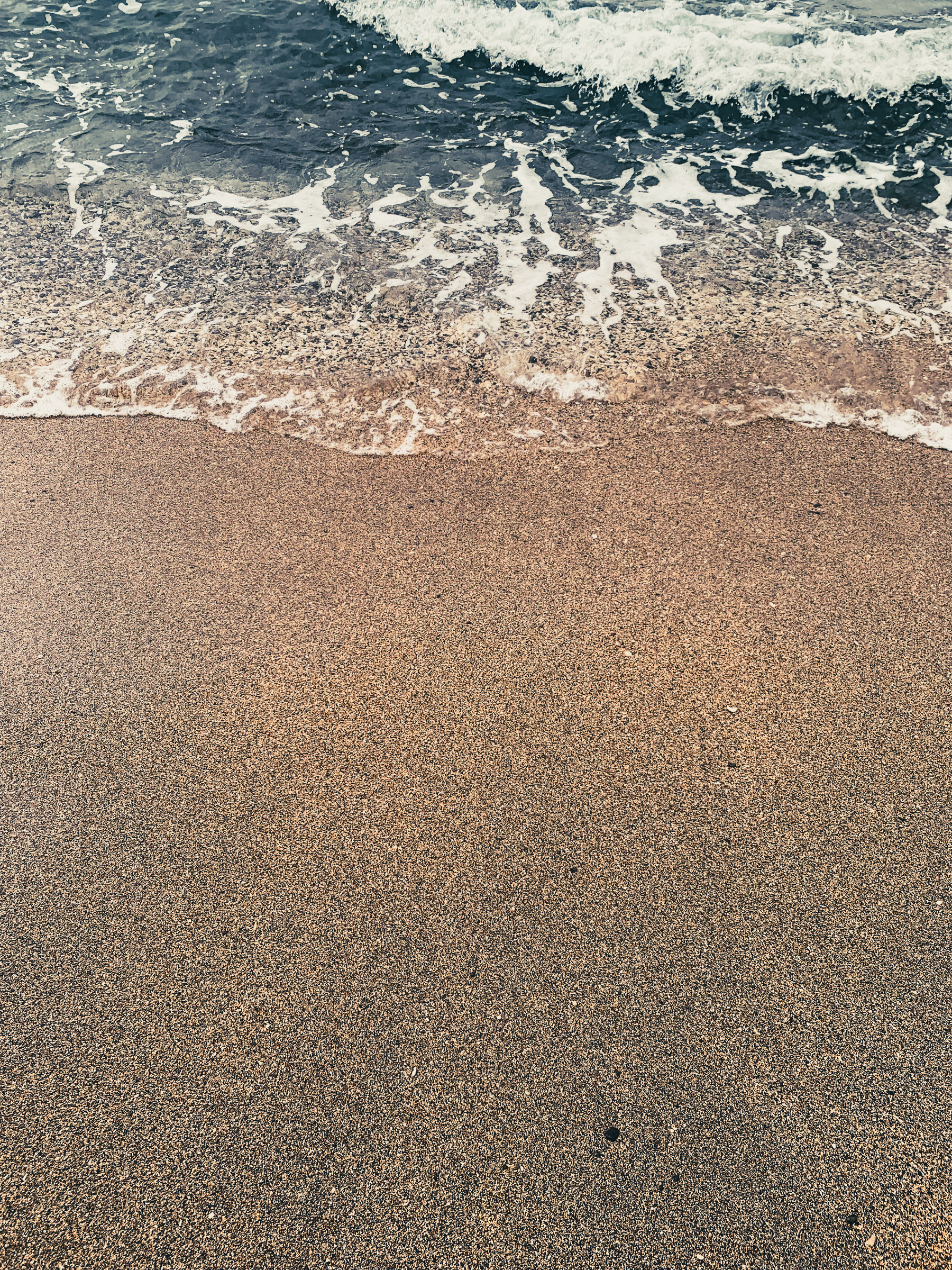 photo of water and sand at the beach. Water fills the upper third of the image, blue-ish green with some ribbony ripples that look like lace, and brown and black smoothly speckled sand fills the lower two-thirds of the image, with a small air hole or two near the lower edge.