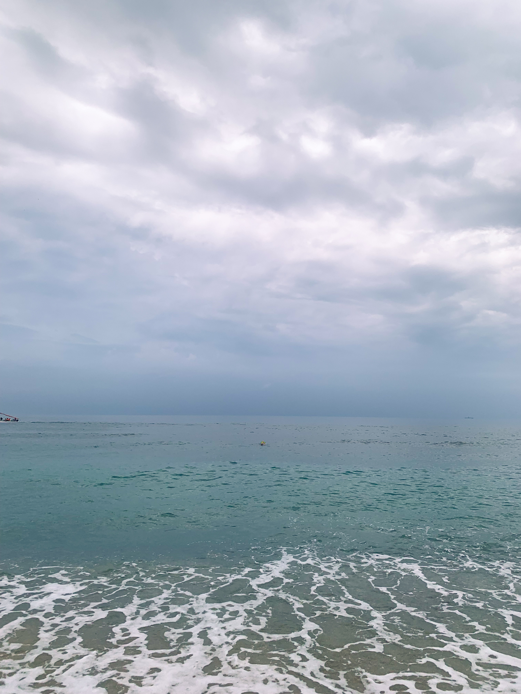 photo of the sea, the horizon, the cloudy sky, with a small boat off to the left edge of the image with a few people in it, one tiny person in neon snorkel gear in the center of the image, and a tiny dim silhouette of a boat to the right of the image, near the horizon
