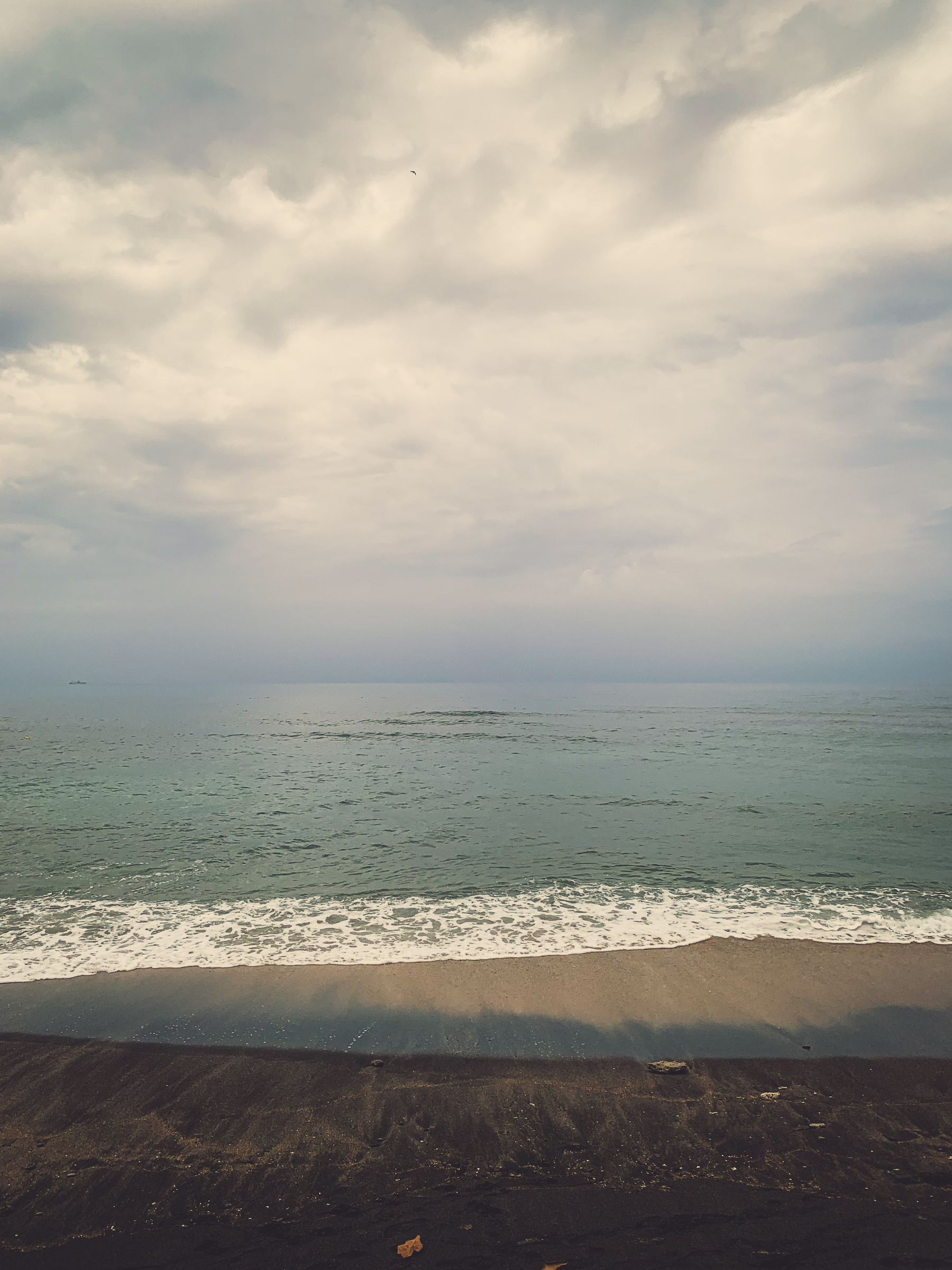 photo of the beach in shifting layers of color, deep brown speckled sand at the bottom with a few old leaves and pieces of coral, strewn with coral gravel, a layer of bronze-beige sand swept by the water, warm greenish to bluish haze of water reflecting light across a disappearing horizon, almost lavender possible rain in the far distance, thick patchy grey clouds over waning yellowish light.