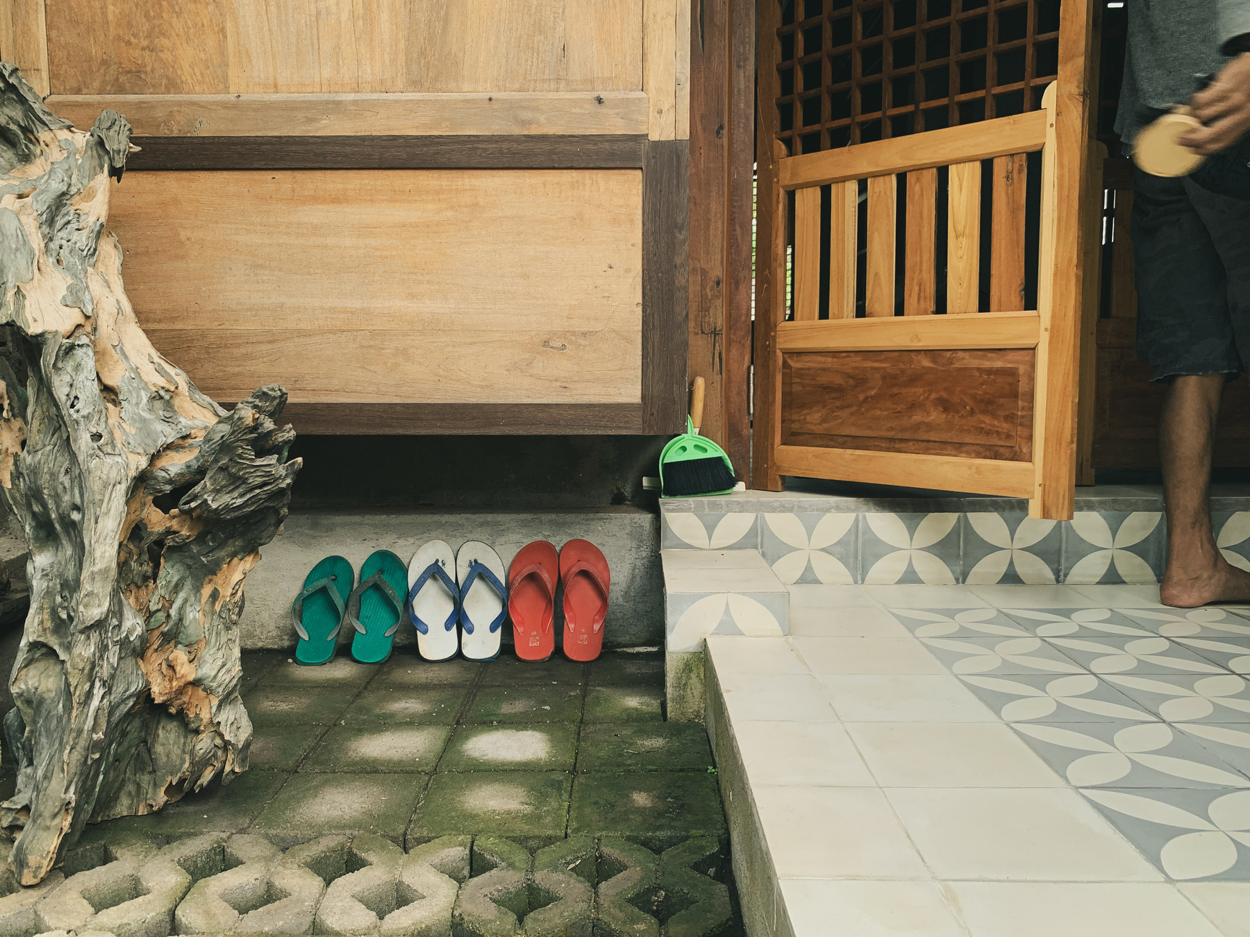 photo of some teak- and ironwood architecture, like a house, with a wood screen door partially open on the right, the photographer’s husband barely in frame standing holding it open, holding also something circular in his hand. The floor is white and grey concrete tiles in a simple kawung motif, and grey concrete pavers, damp and slightly mossy, with an interesting, elaborate, aged piece of wood to the left of the image, and three pairs of well-worn sandals, brightly colored in red, white/blue, and green, leaned up against the wall, in the left side of the image, with a bright lime green small dust pan and broom leaning next to the door.