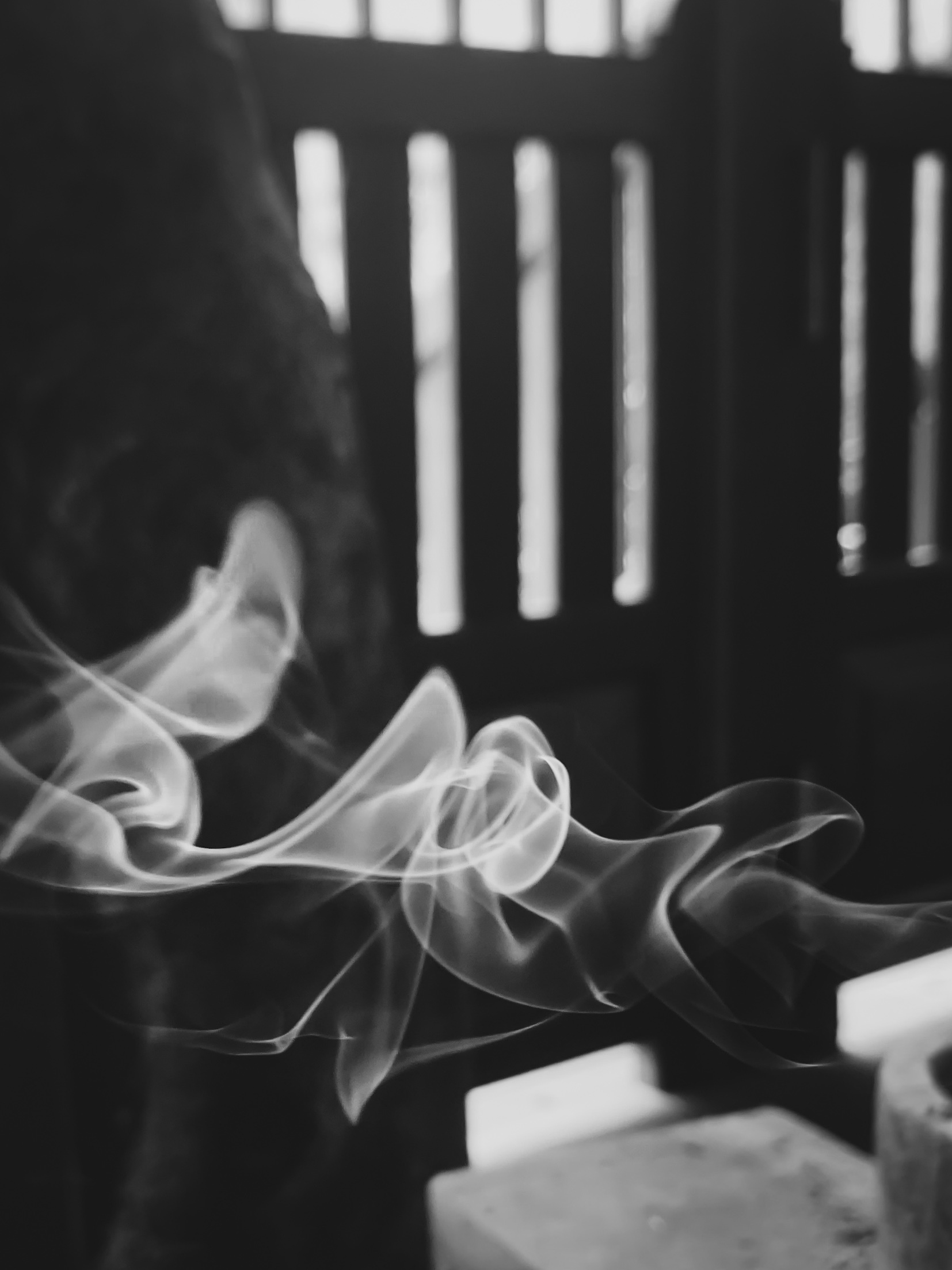 black and white photo of white incense smoke making sinuous and ghosty shapes across the lower third of the image, with blurry slatted woodwork in the background.