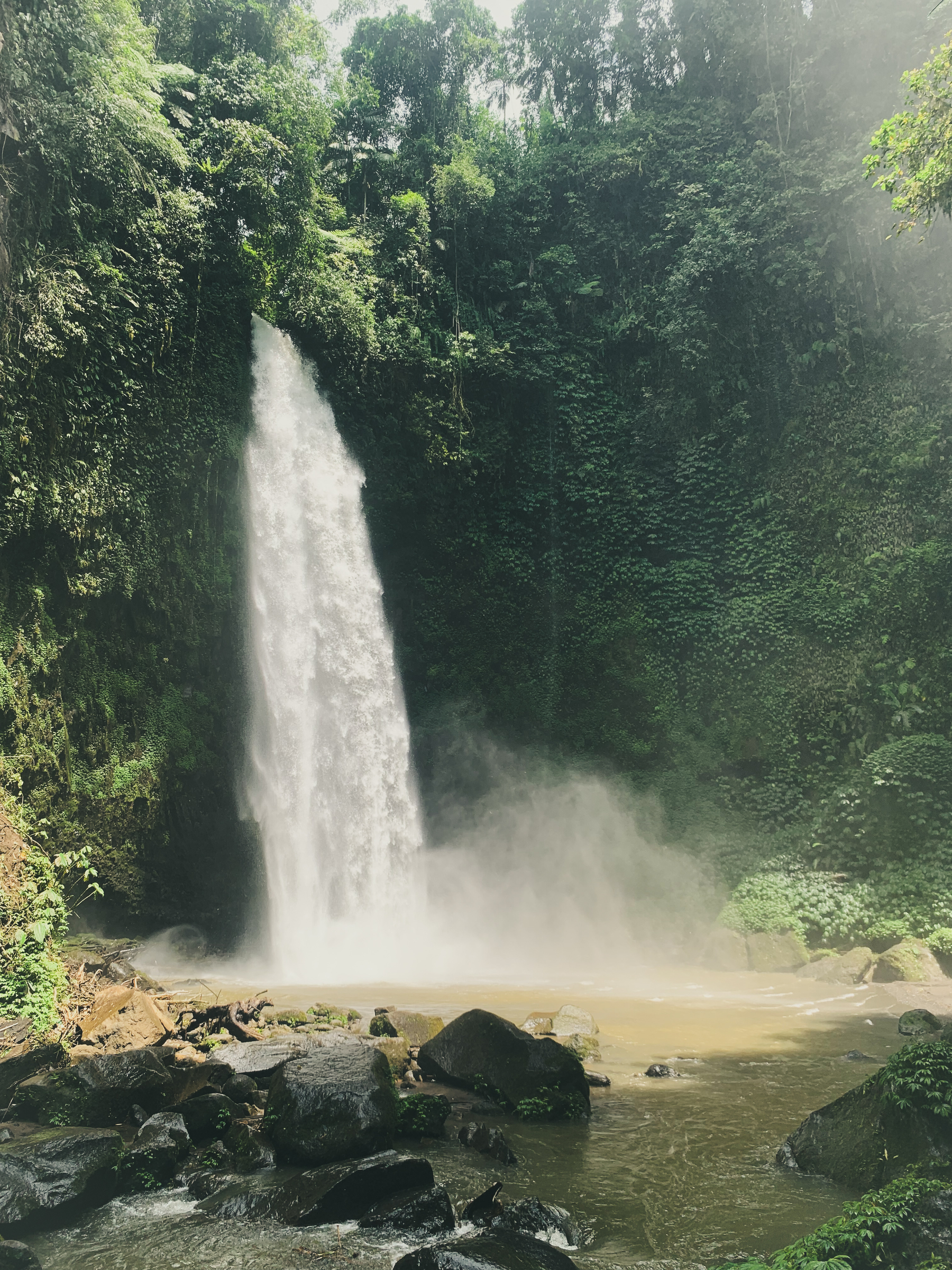 photo of a waterfall catching sunlight surrounded by lush green tropical forest emptying into a brownish pool and throwing up a gusty cloud of mist and flowing down around and over boulders and rocks.
