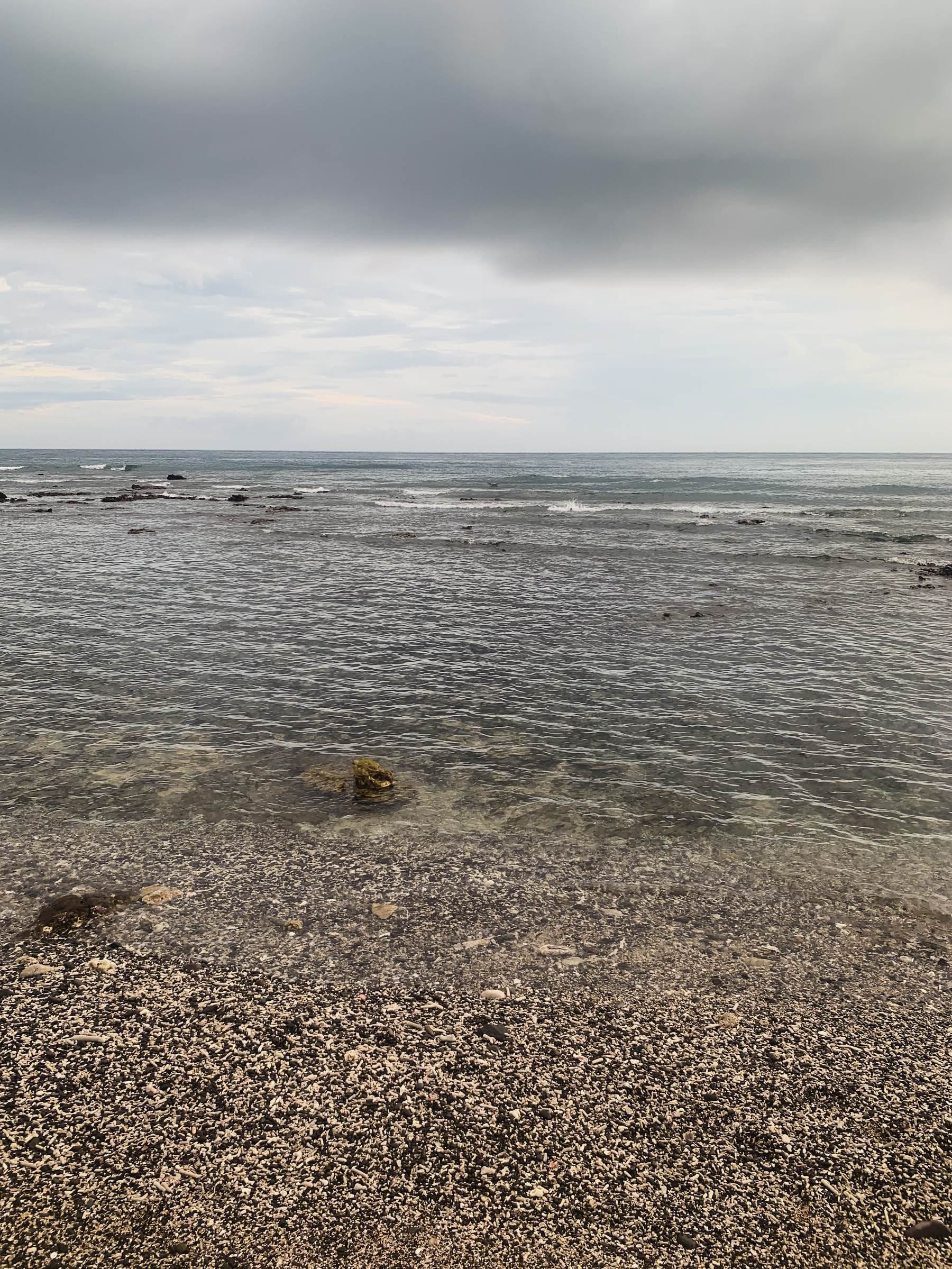 photo looking out to sea from the beach with a mass of grey cloud over a clearer sky, small breakers at the reef on not too wavy but rippled grey-green-blue water, and speckled tan and white and black gravel and coral pieces and sand