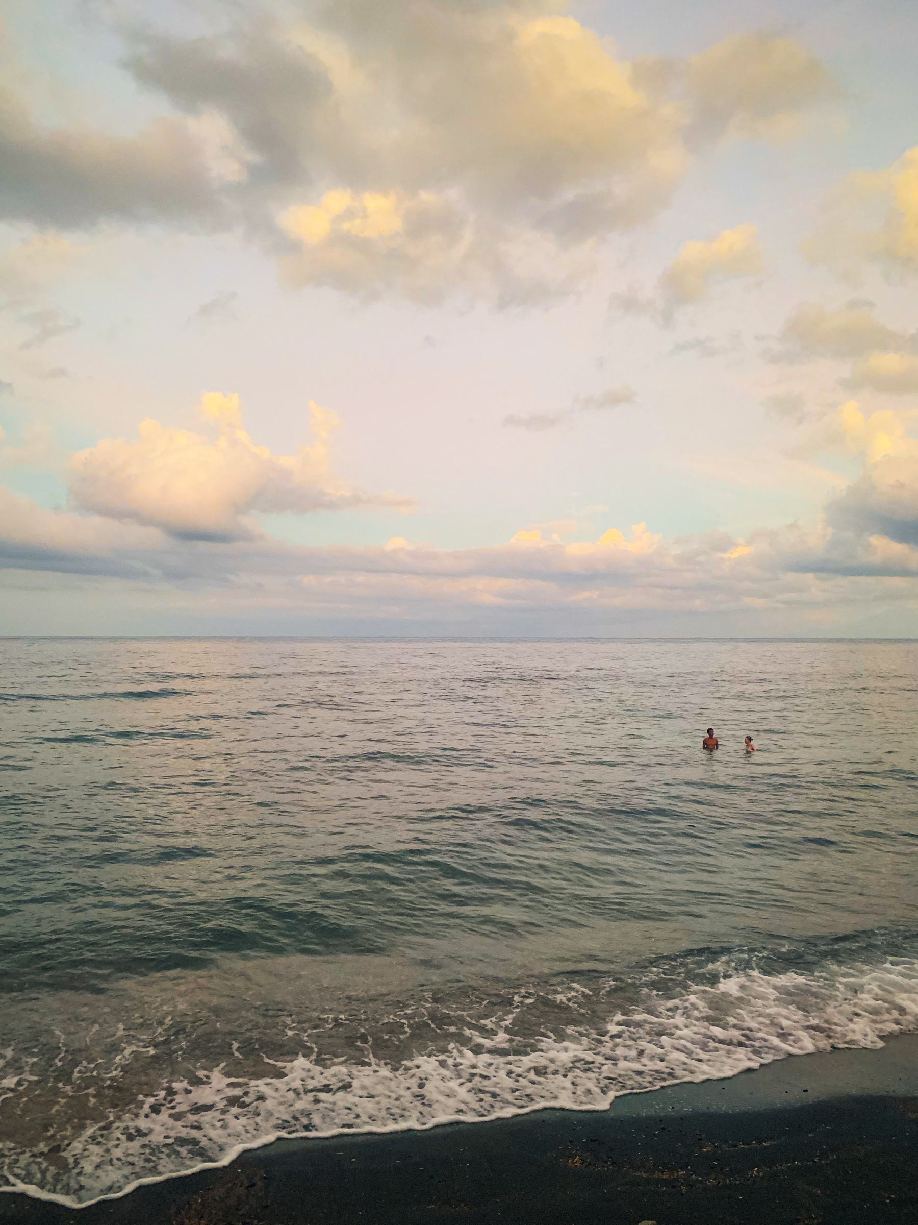 photo at the beach looking out to the sea at sunset with pale pastel pink, blue, yellow and grey clouds and calm but wavy water washing up onto black sand and two people in the water off-center in the distance