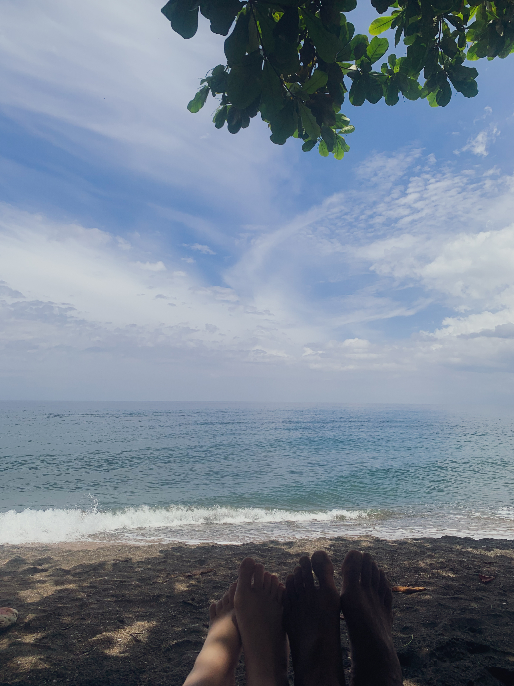photo of two peoples feet in the shade looking out at a beach, with blue sky and wisps of white clouds, and green leaves hanging down from above.