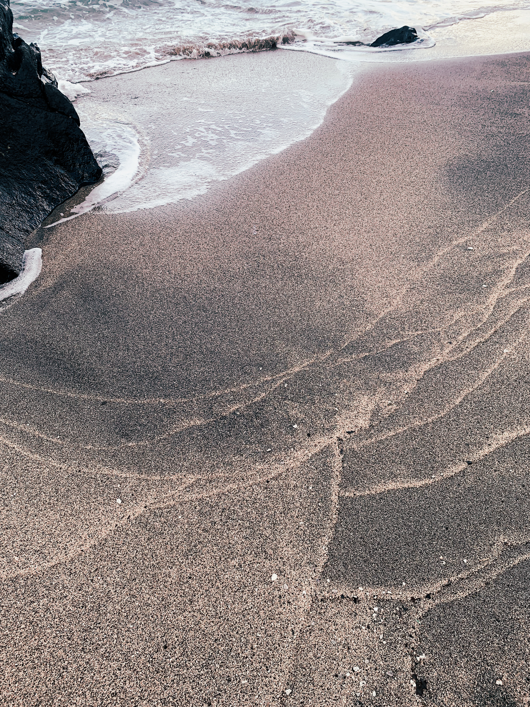 photo at the beach where waves have made contoured patterns in beige and black sand, with foamy sea water moving around a volcanic rock in the upper left, and bright warm sunlight reflecting off of water in the upper right corner of the image.