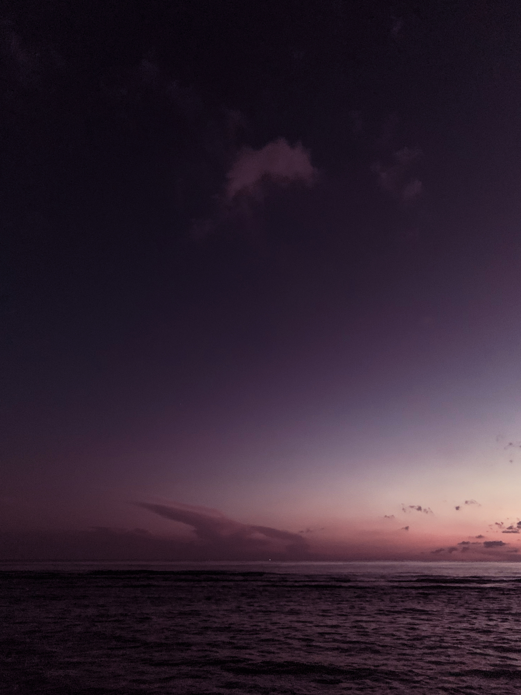 photo of the sea reflected dark purple under dark purple sky of pre-dawn with a rose-colored glow across the horizon and some unusual cloud formations with a tiny speck of light at the center of the horizon