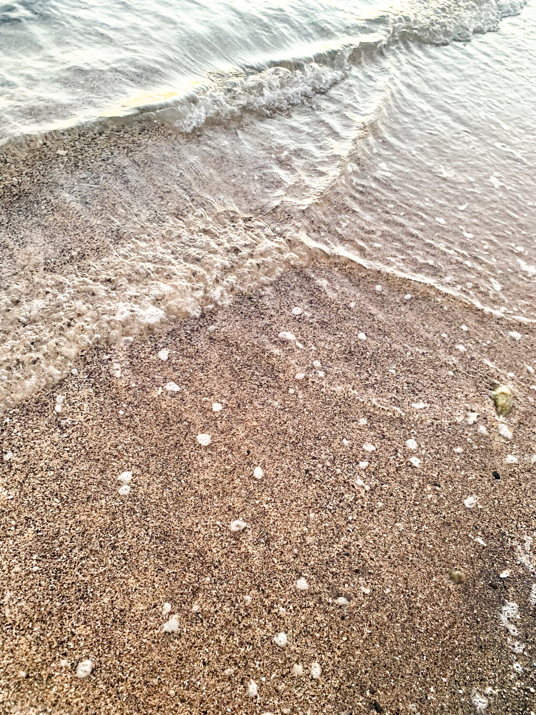 photo at the beach of clear sea water washing in a sexy ripple over beige sand with black speckes in it and small touches of foam.