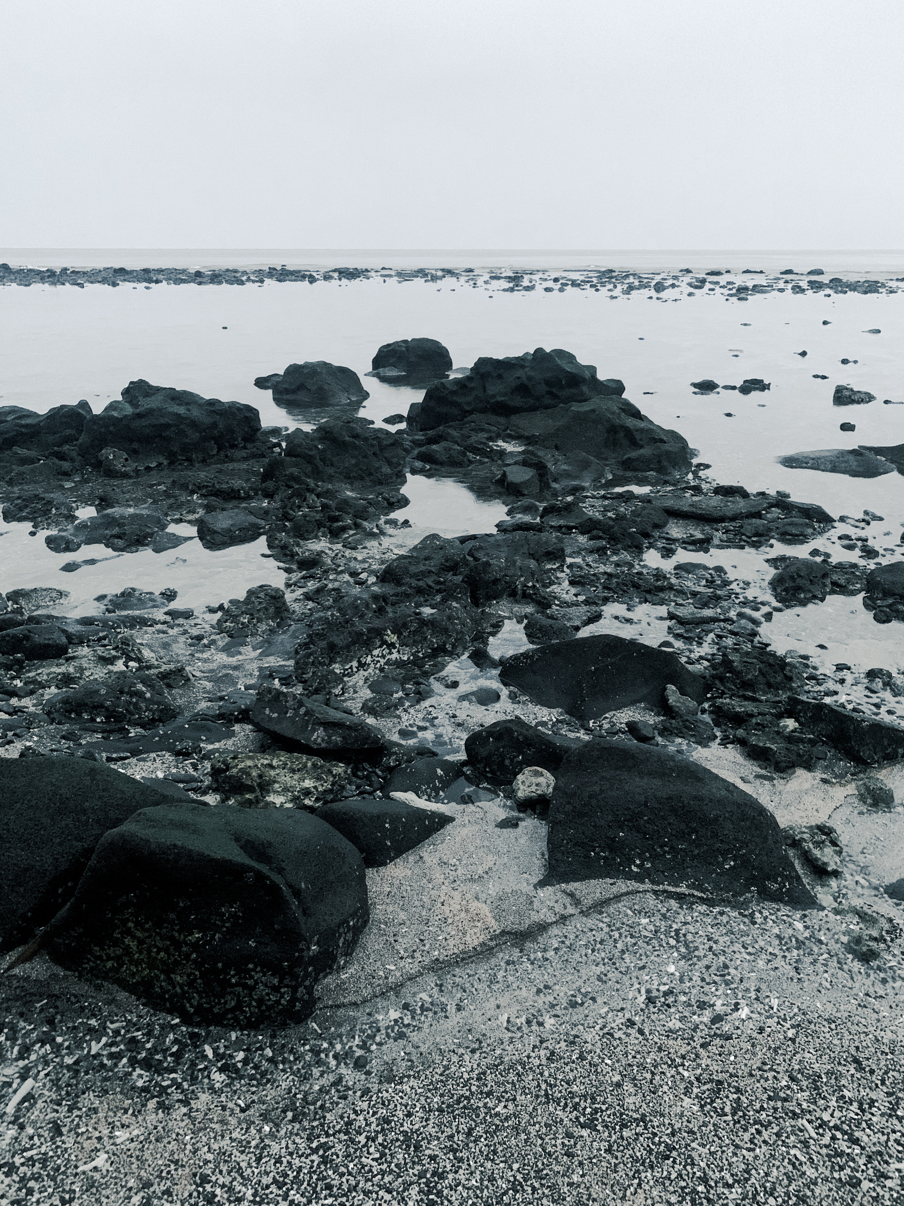 a dramatic black and white photo taken at the beach of black volcanic boulders sitting at the shoreline, at low tide, with nearly still water and dead coral reef in the distance.