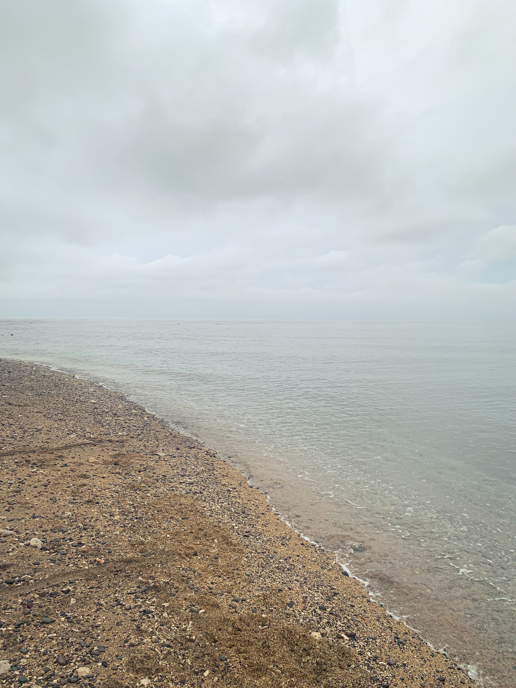 photo at the beach of very calm water, meeting beige gravel- and pebble-strewn sand in a line that curves off to the left, with the water surface reflecting the grey hazily clouded sky, making the horizon nearly invisible.