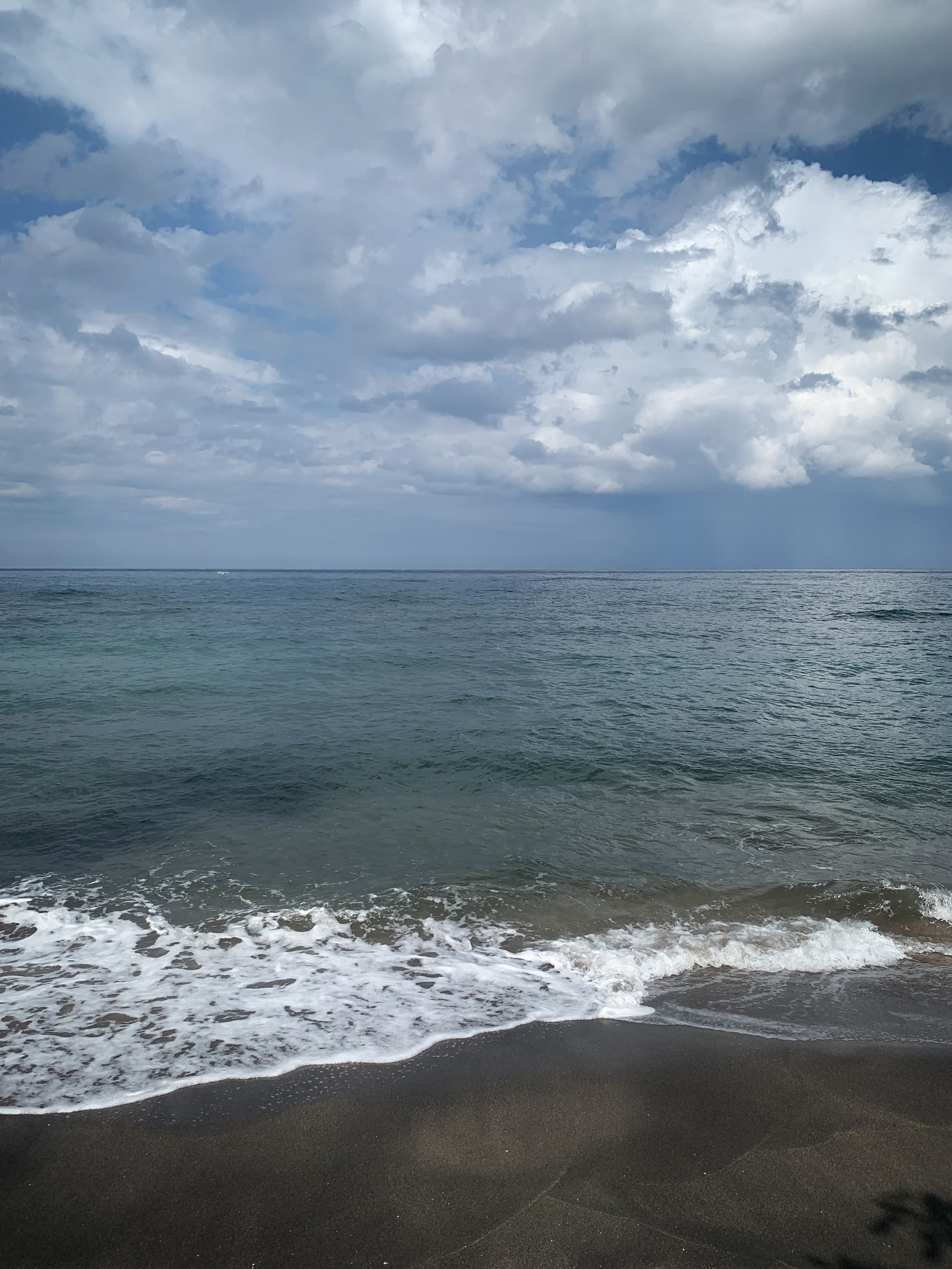 photo of the beach with calm green-blue water frothing up onto brownish black sand, the horizon near the middle of the image, and the sky blue and full of lofty white clouds, and maybe a rainstorm in the distance.