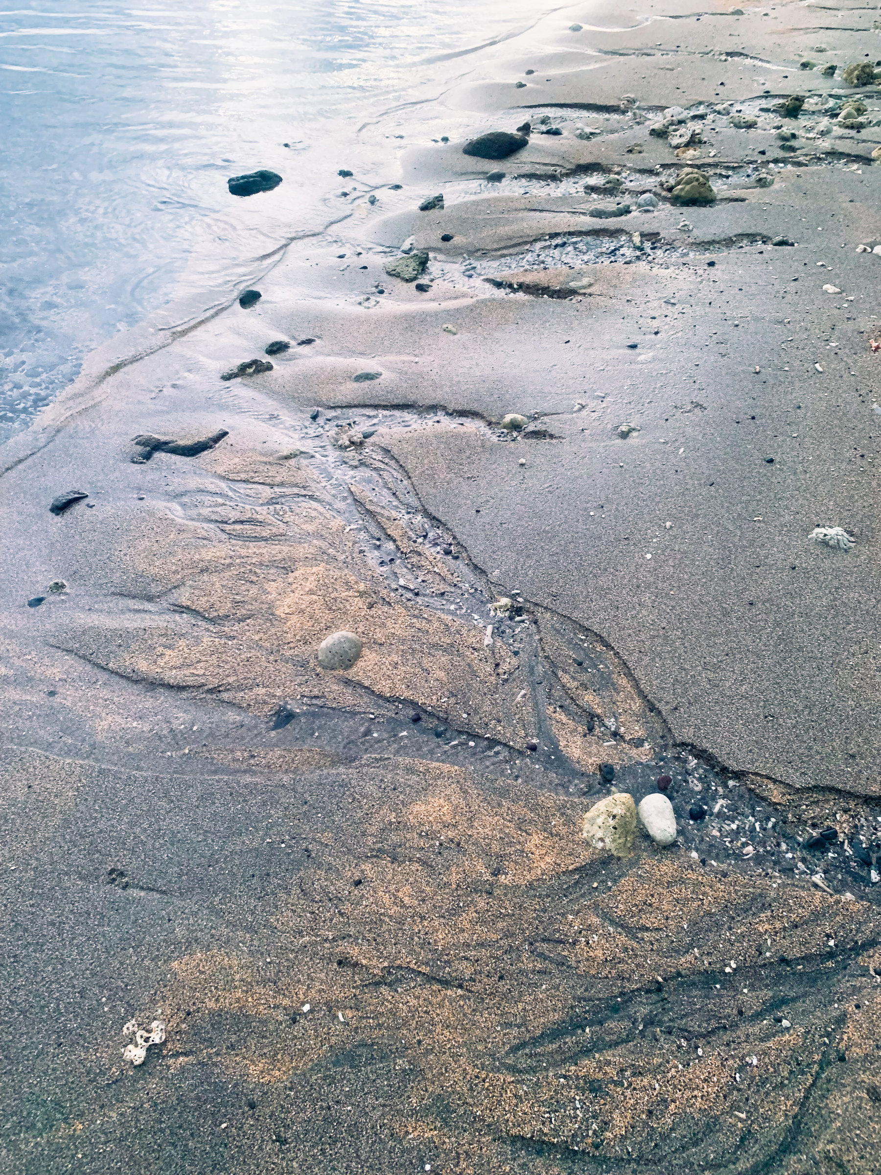 photo at the beach of the edge of the water, with small streams of water trickling back through the beige and grey sand down into the low tide, which is in the upper left corner of the image; with water carving many feathered and contoured ways into the clear water, reflecting pale and silvery sunlight, studded with occasional black and grey volcanic rocks or chunks of coral, and smaller stones and pieces of shells.
