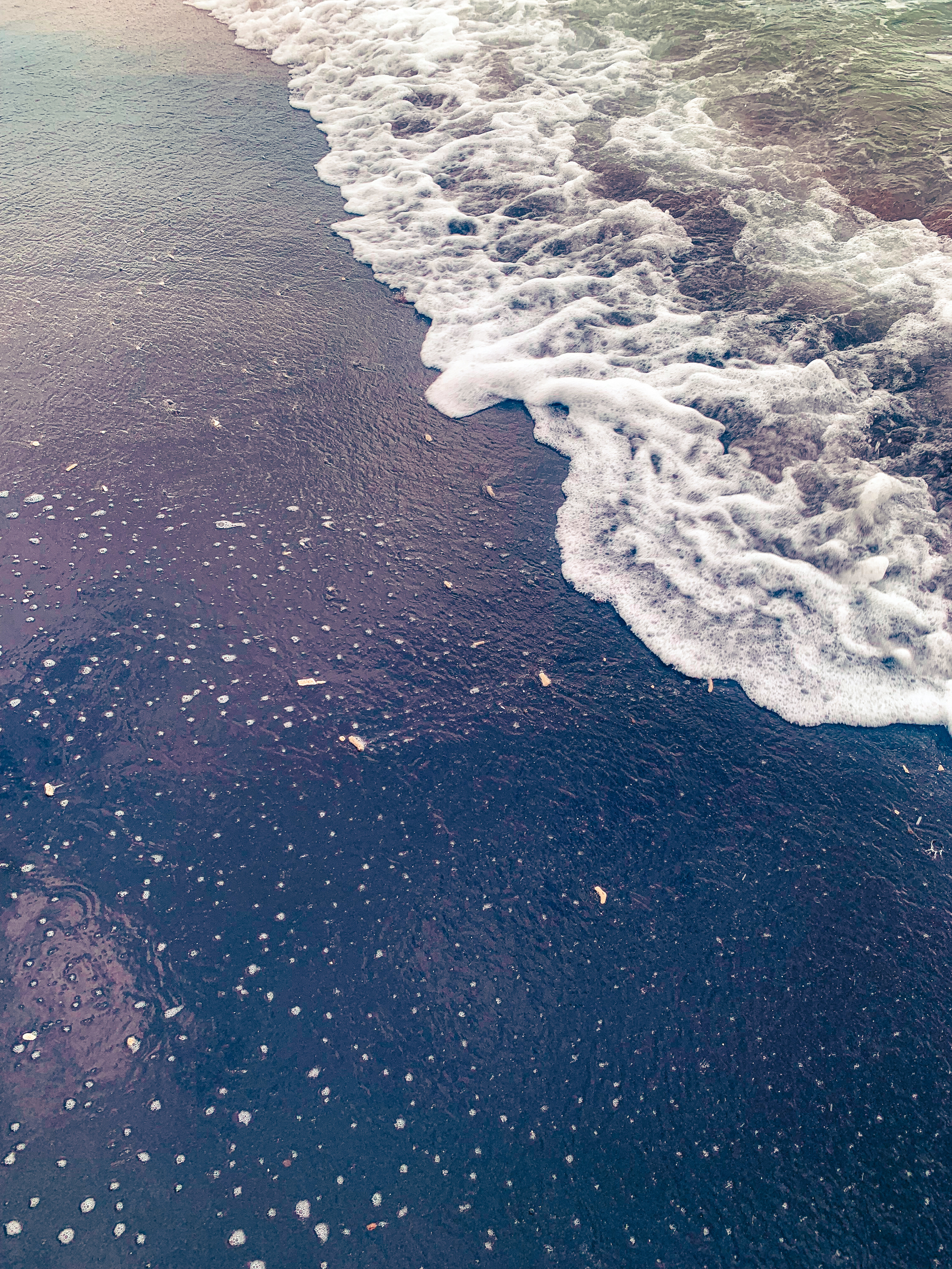 photo is at the beach at the water’s edge, with frothy water churning in the upper right portion of the image, with a oily film or glasslike water spread and rippling across black sand, with dots of froth, filtering and reflecting light in bending shades of greenish, pinkish, and brownish blue-black.