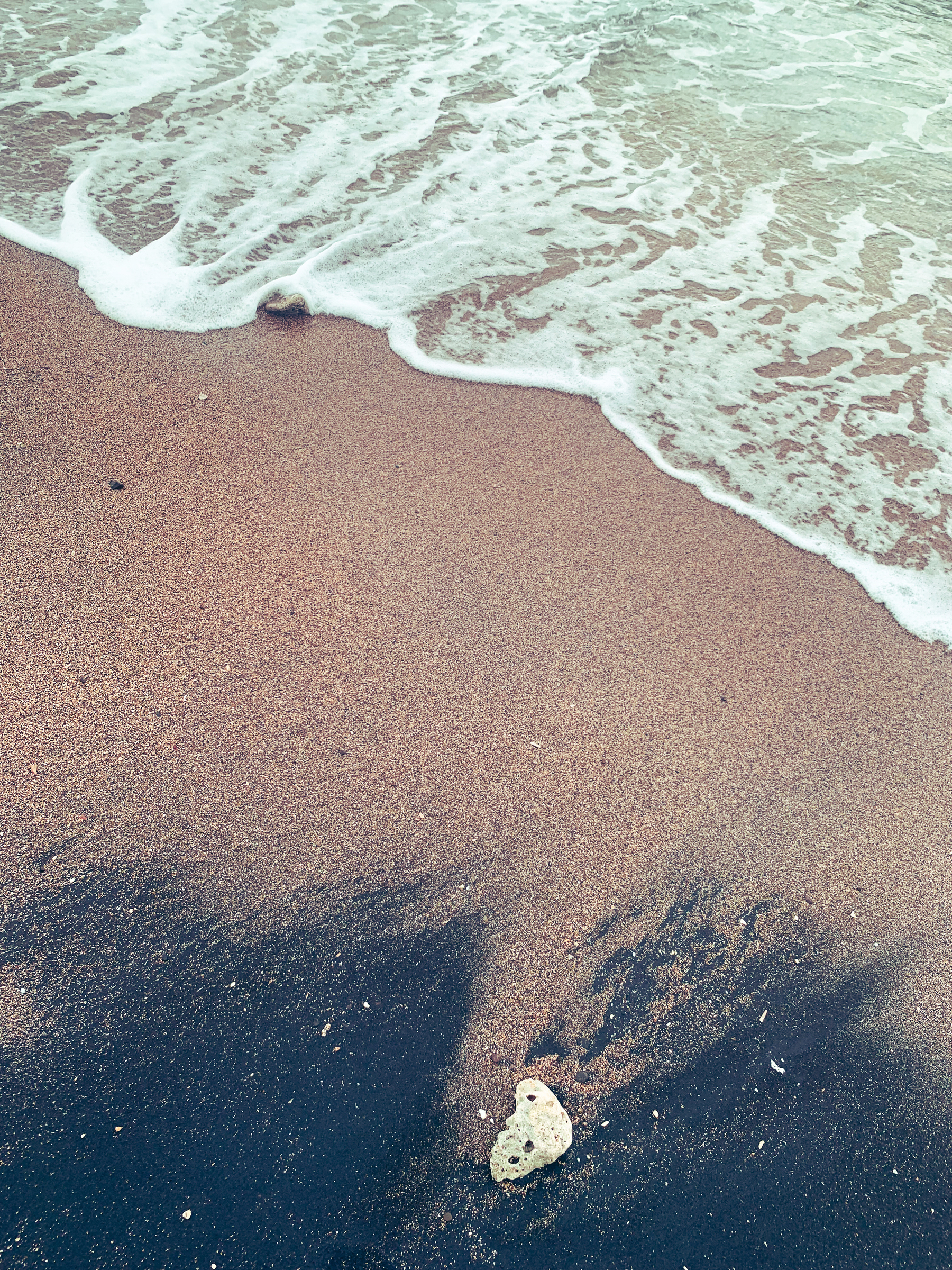 photo at the beach of frothy sea-green water washing up on a pinkish tan sandy shore with submerged pieces of coral in it and one is being touched by the water