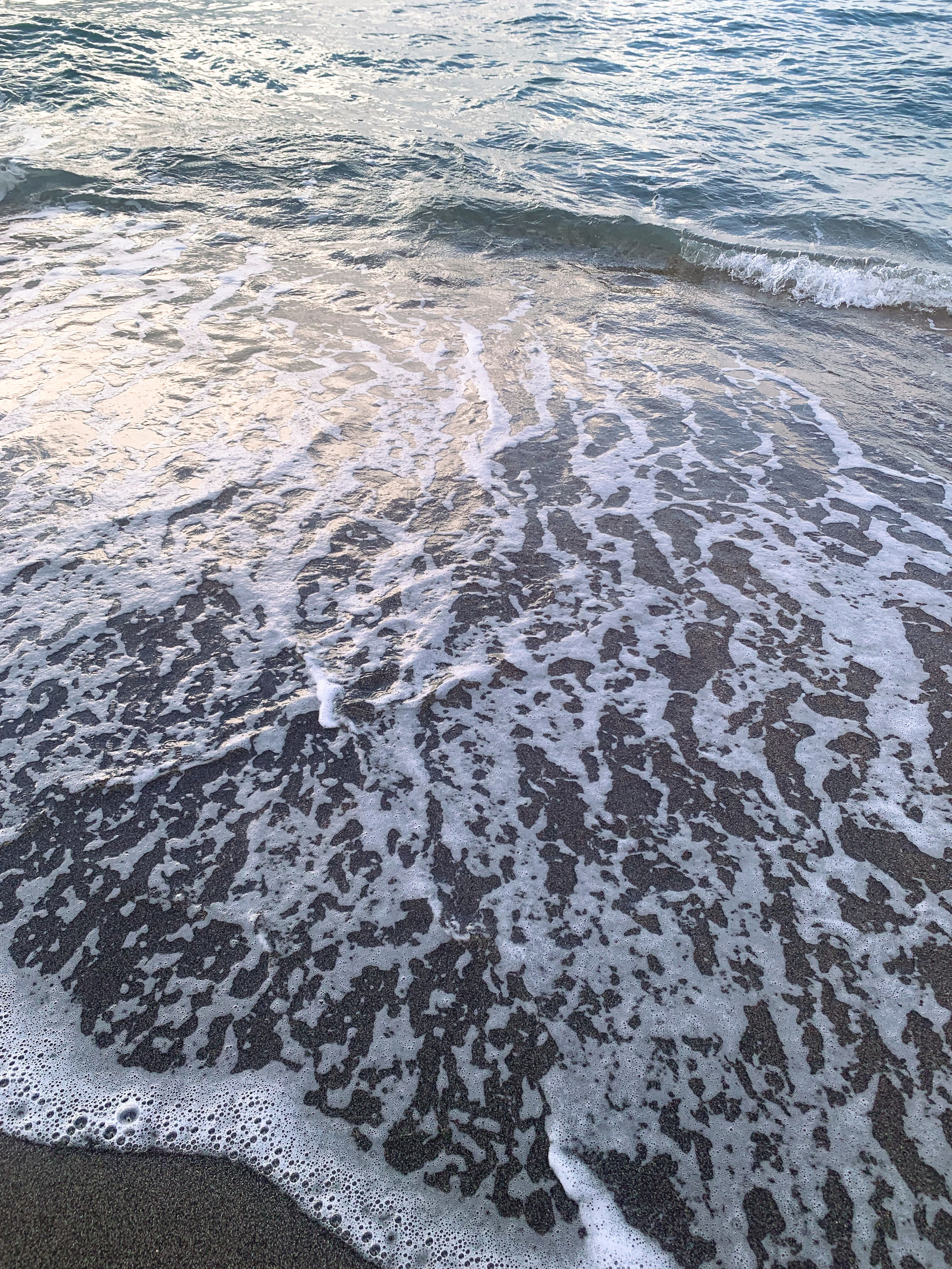photo of water at the beach skimming over beige-grey sand with froth and bubbles, reflecting sunlight almost like metal
