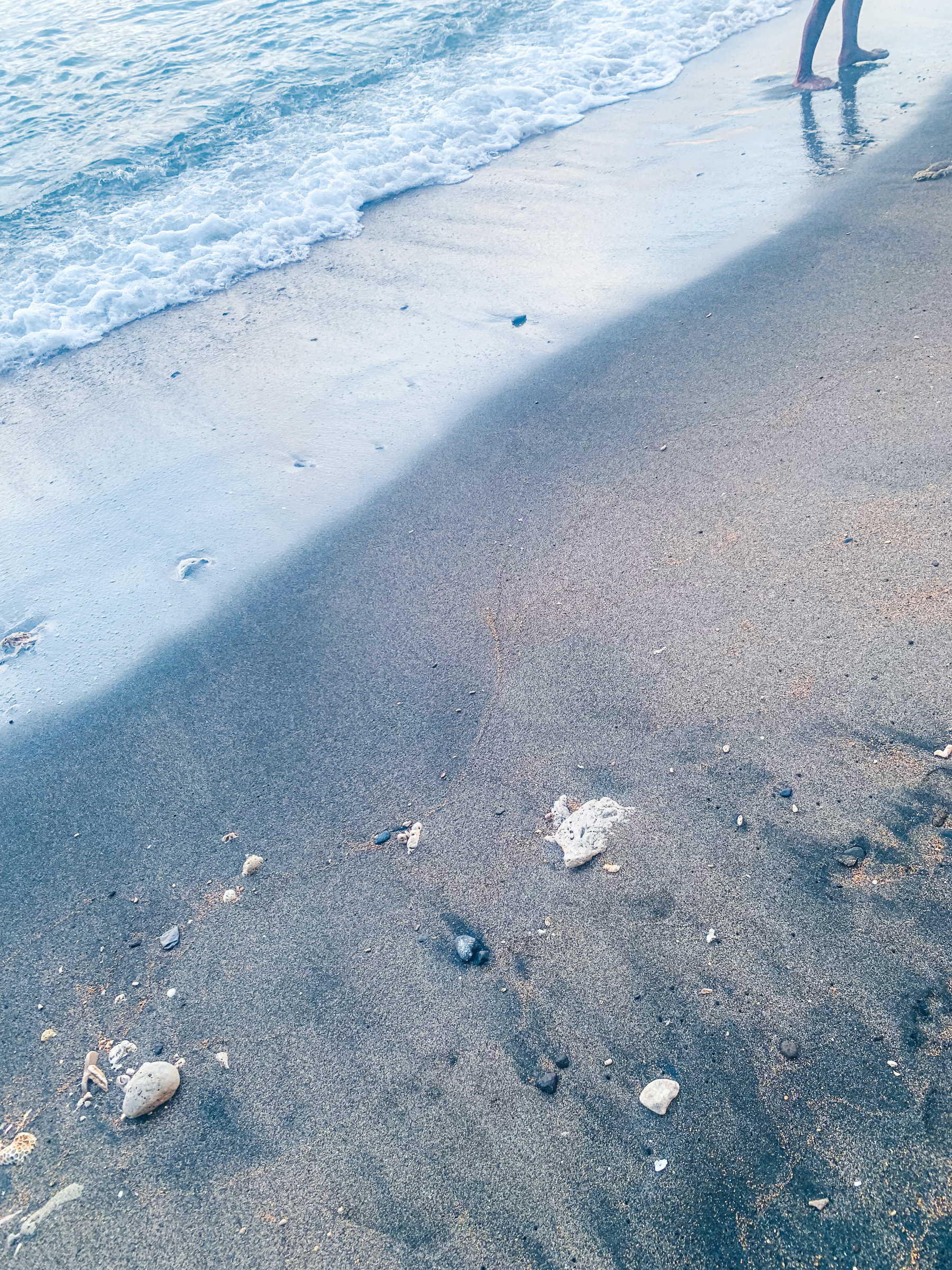 photo at the beach with pale blue misty light of the shoreline washing onto beige sand with some drifted chunks of coral and shells. a person’s lower legs and feet are visible in the upper right corner of the image and a few footprints faded into wet sand
