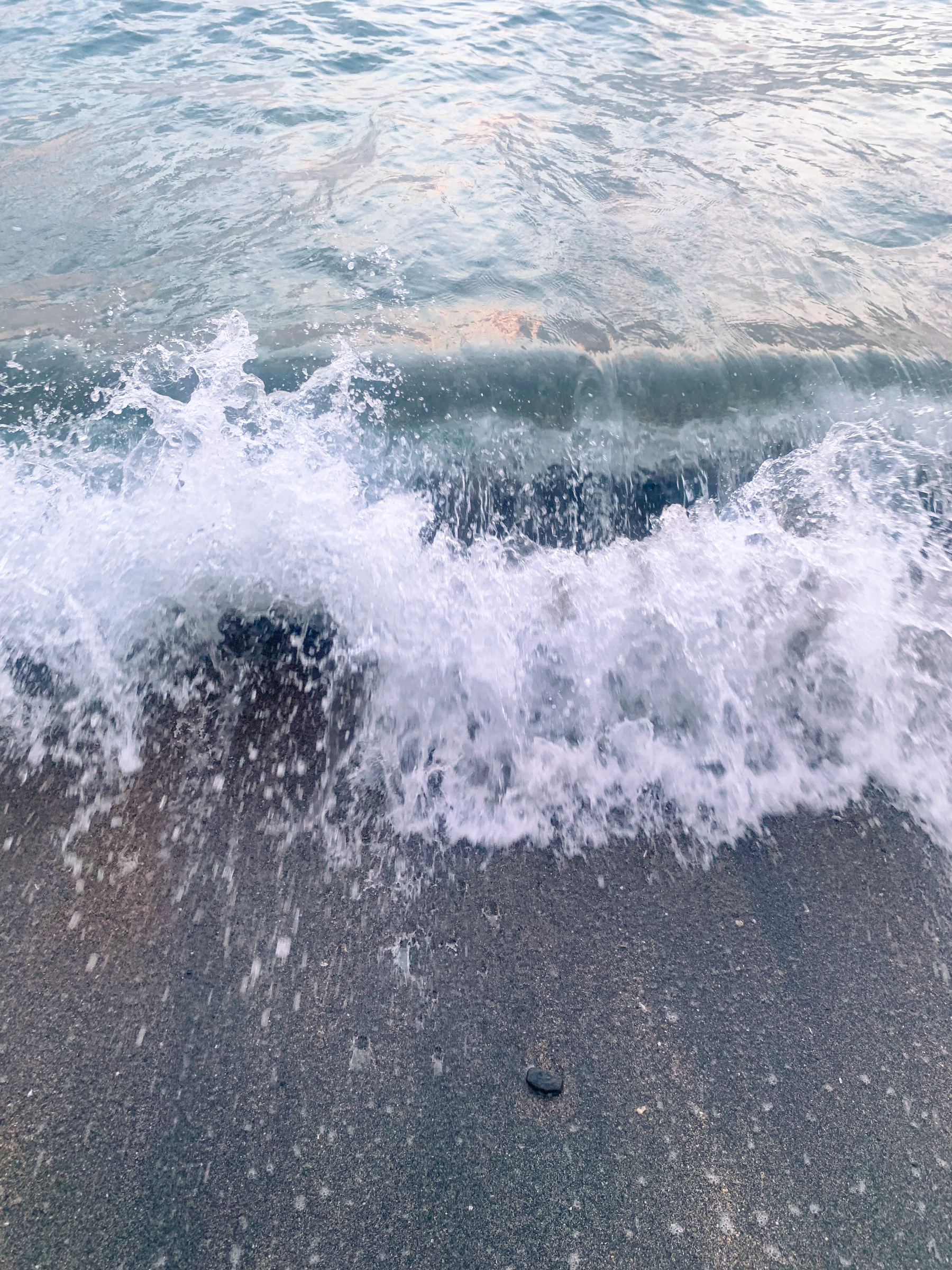 photo at the beach of a small wave splashing at the viewer, with sea-green water and blackish-tan sand.